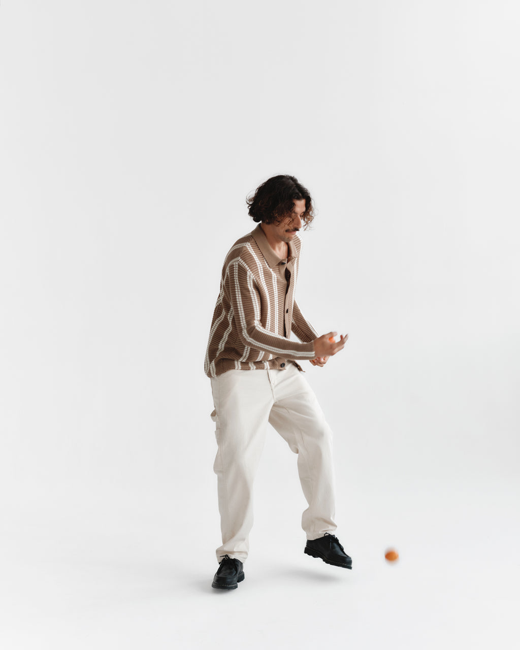 A person with wavy hair is juggling a small ball while wearing the Dezz Stripes LS - Brown Stripe by Gabba, paired with white cotton pants and black shoes, set against a plain white background.