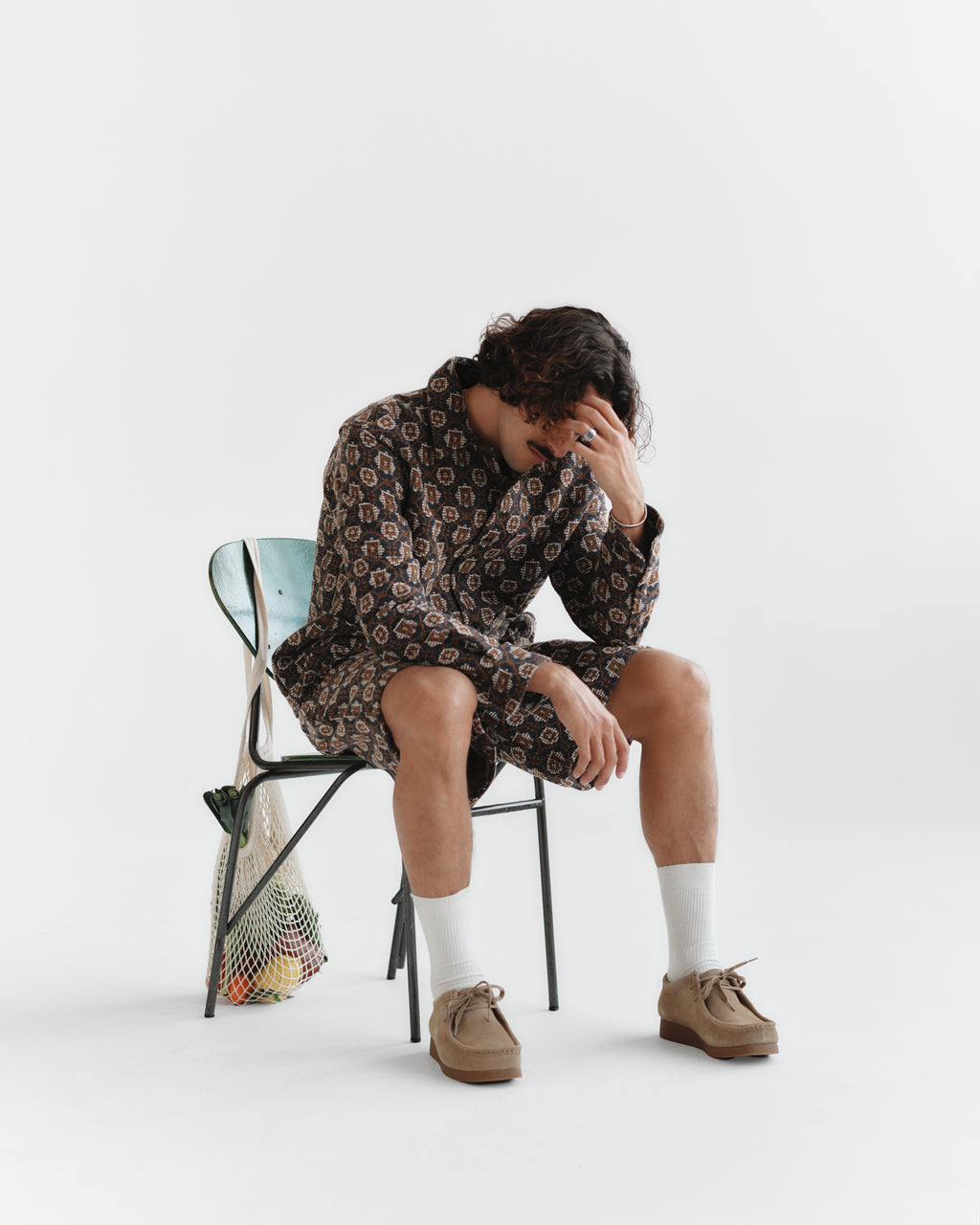 Someone sits on a chair in Gabbas Fede Box Star - Multi Pattern outfit, showcasing boxy shorts and a long-sleeved top. They rest their head on their hand, with a net bag hanging from the chair against a white background.