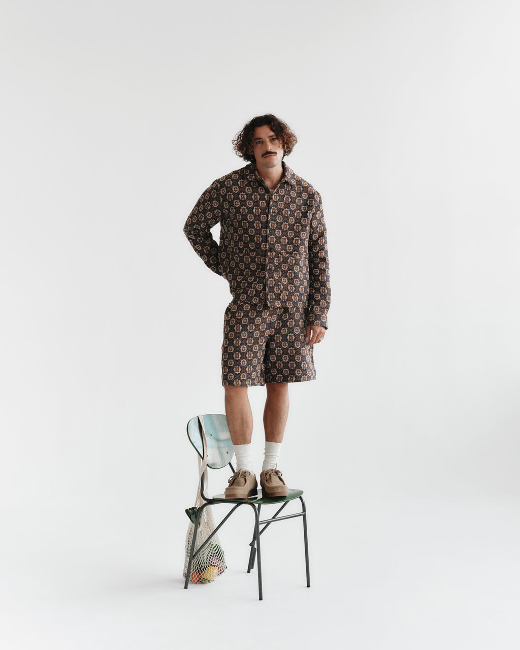 A curly-haired person stands confidently on a chair against a plain background, wearing Gabbas Fede Box Star Multi Pattern shorts and shirt set. Their hands rest casually in their pockets, while a mesh bag with fruit hangs from the chair back, completing the ensemble.