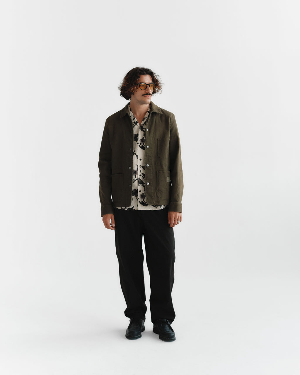 A person with curly hair and glasses stands against a plain background wearing a dark jacket over the Gabba Sven Reg Resort Bloom SS - Meteorite shirt paired with black pants and shoes. Their look is casual yet stylish.