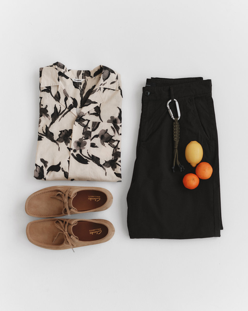 A flat lay features the Sven Reg Resort Bloom SS - Meteorite by Gabba, dark pants with a carabiner, brown suede shoes, and three fruits (a lemon and two oranges) on a white background.