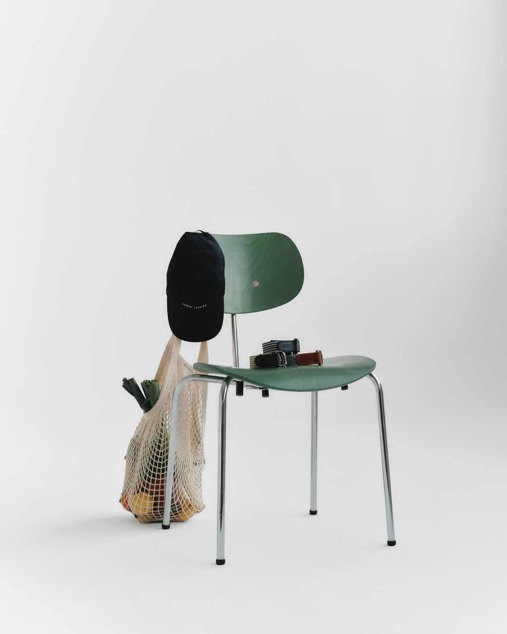 A green chair with chrome legs sits on a white background. On the seat are a black camera and sunglasses. Hanging from the backrest: a black cap with Gabba’s Manica Belt – Green Stripe, plus a mesh grocery bag of produce.