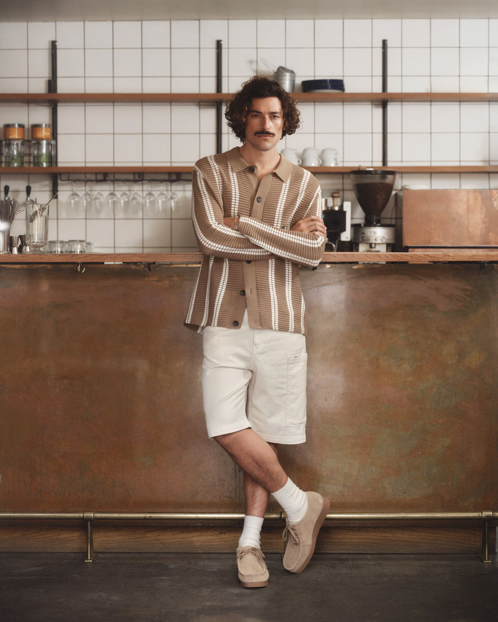 A curly-haired, mustached individual stands confidently before a trendy bar, wearing a beige and white striped shirt paired with Gabbas Maxwell Luis Cargo Shorts in Ecru. Their non-stretch outfit is finished with socks and beige shoes, set against shelves filled with glassware and jars.