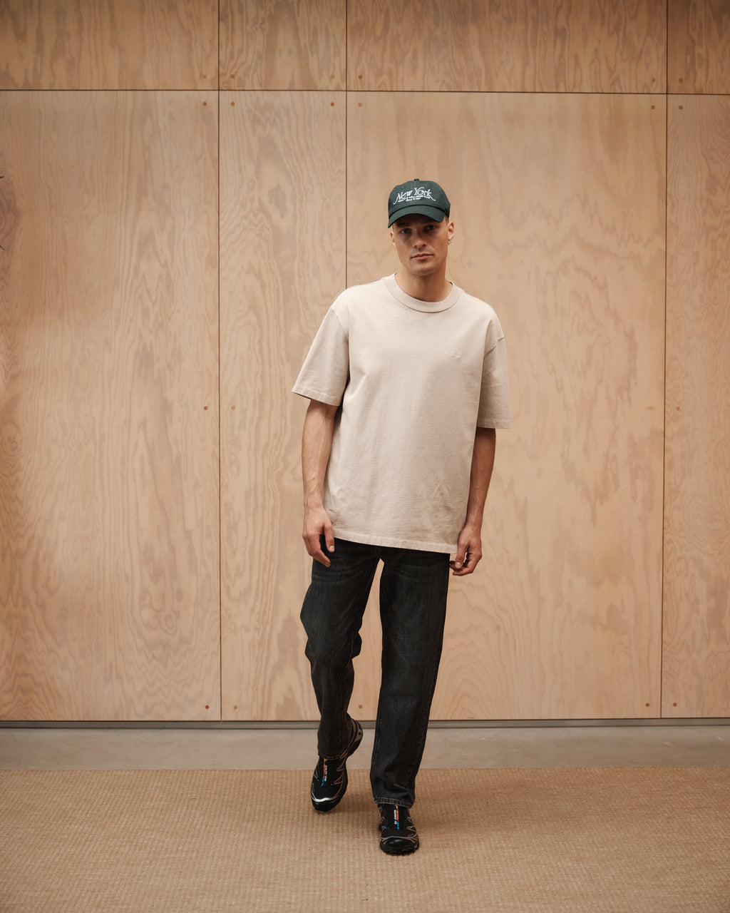 A man in a beige oversized t-shirt, Gabba Max F1011 - Black Denim jeans, black sneakers, and a dark green cap walks indoors by a wood-paneled wall and light brown carpeted floor.