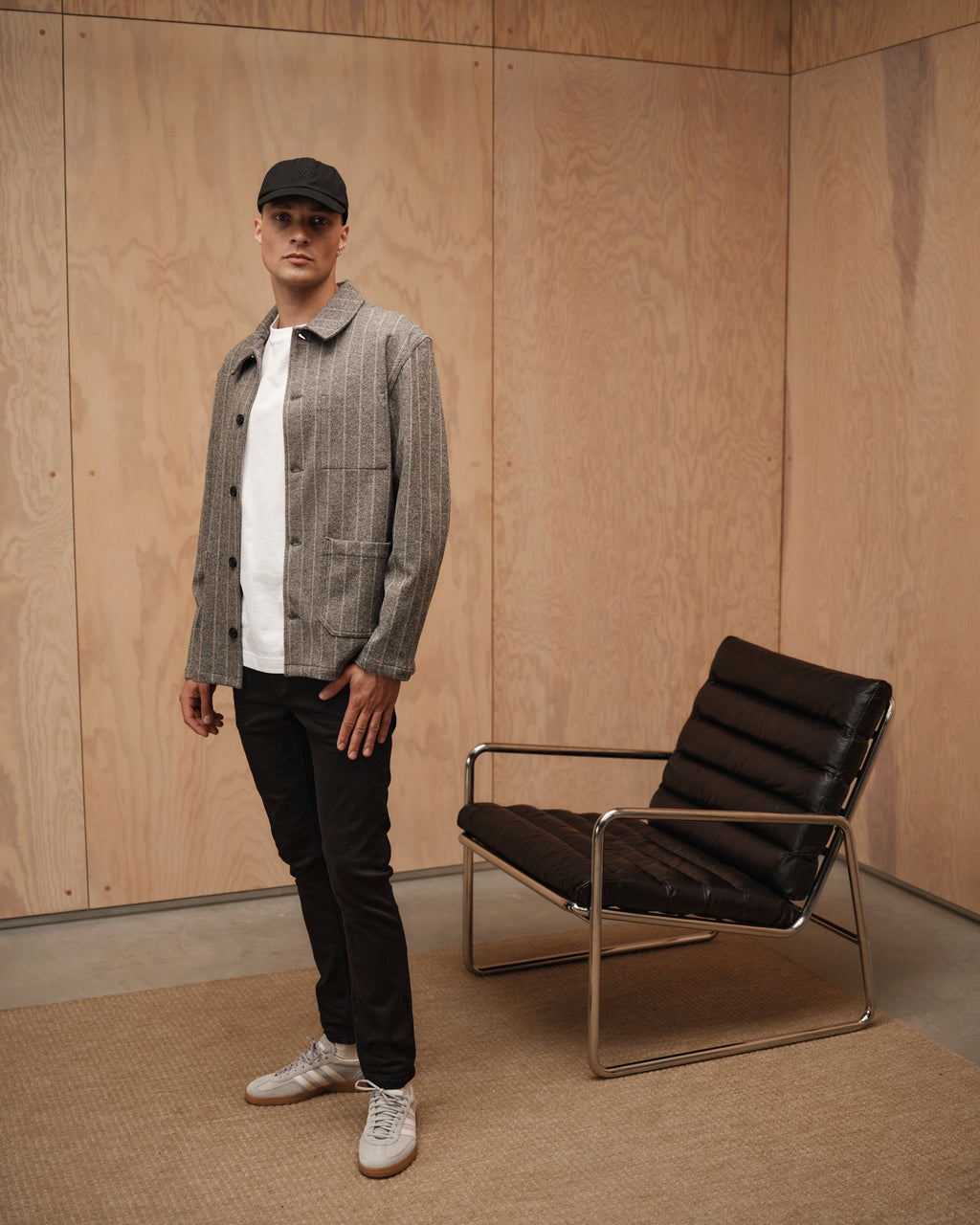 A man wearing Jones Hold Black - Black Denim jeans by Gabba, a gray jacket, white t-shirt, white sneakers, and a black cap stands beside a modern black leather chair in a wood-paneled room.