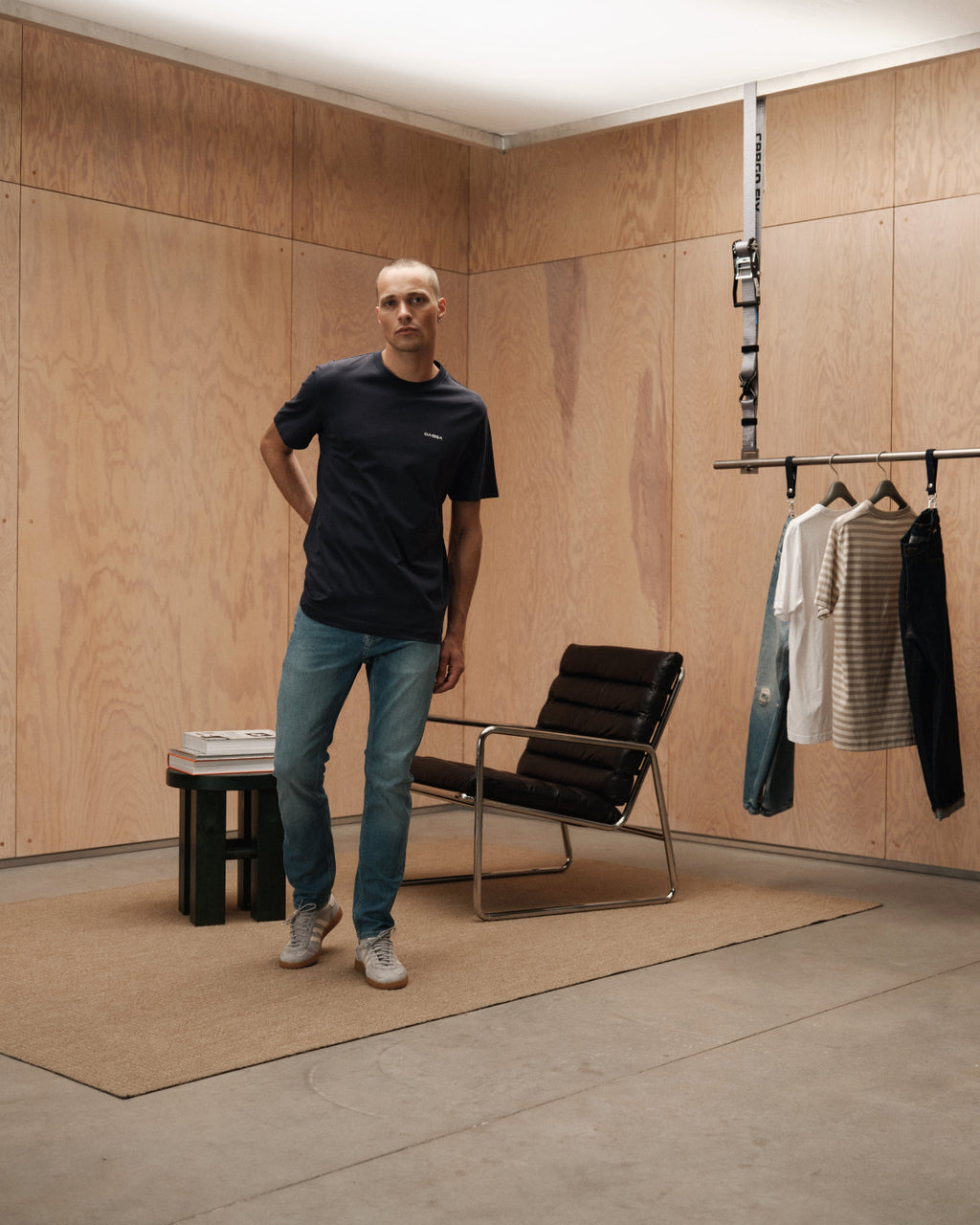 A man in a navy t-shirt and GABBA Jones K2615 Lt. - Denim wash jeans with a slim fit stands in a minimalist room with wood-paneled walls, near a chair, small table with magazines, and a rack holding shirts and pants.
