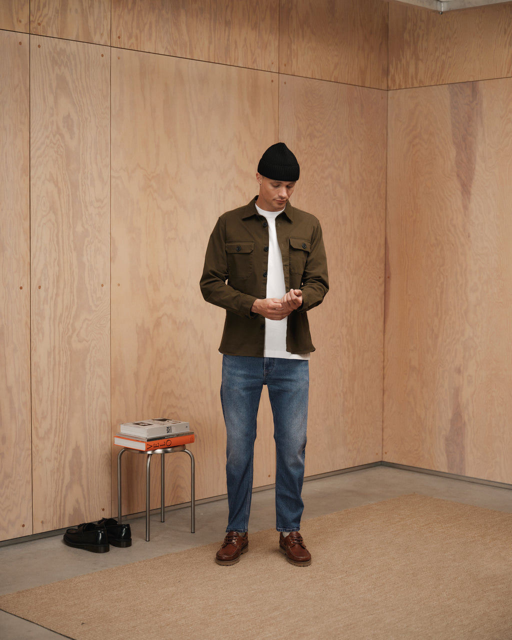 A man in a black beanie, olive green jacket, white shirt, and Gabba Carl Oskin Dark Blue Denim jeans stands on a beige rug in a wood-paneled room. Black shoes complete his casual look; books and glasses rest on a nearby stool.
