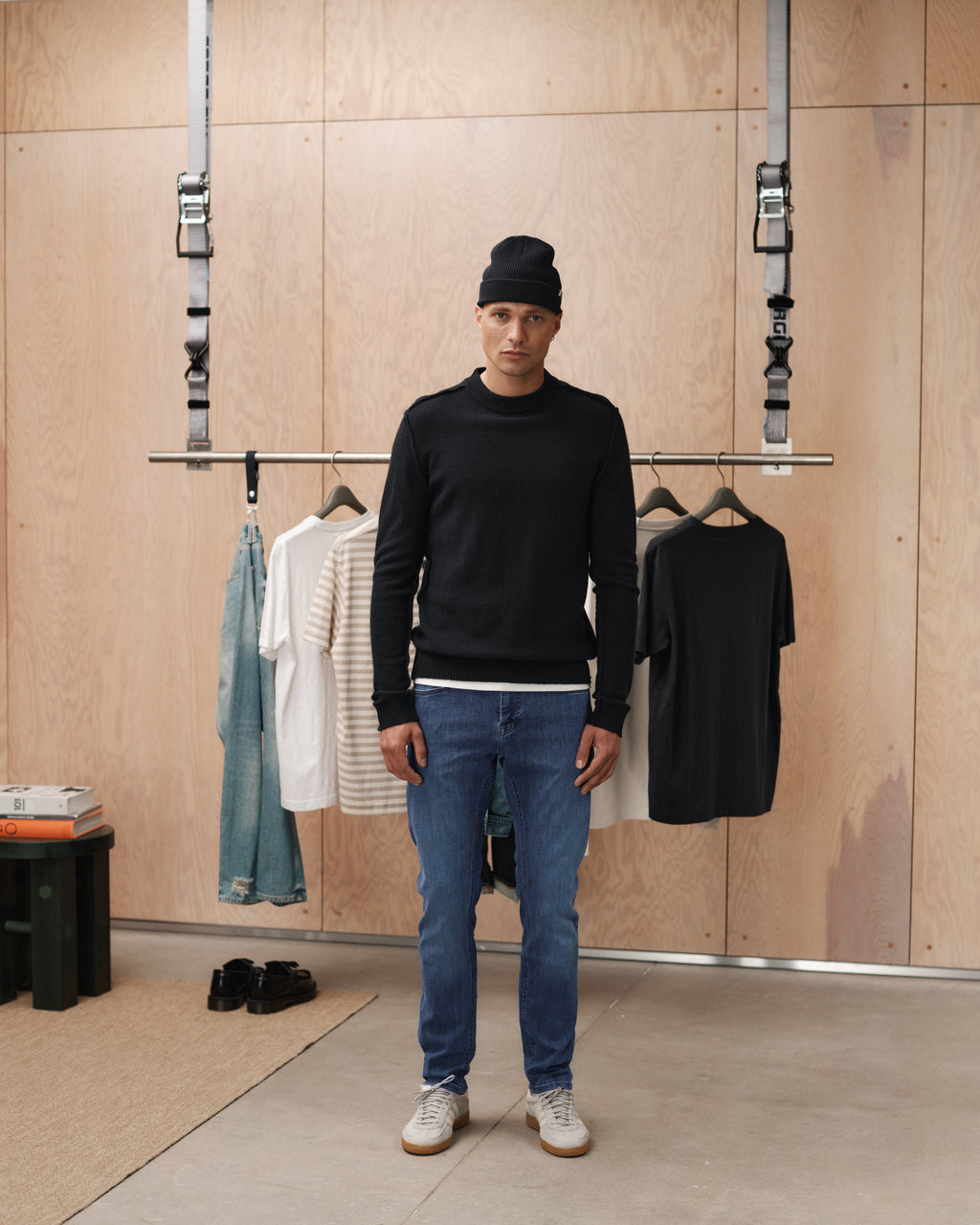 A man wearing a black beanie, black sweater, Jones Vanew - Mid Blue Denim jeans by Gabba, and white sneakers stands indoors before a clothing rack with slim silhouette jeans and shirts against a wooden wall backdrop.