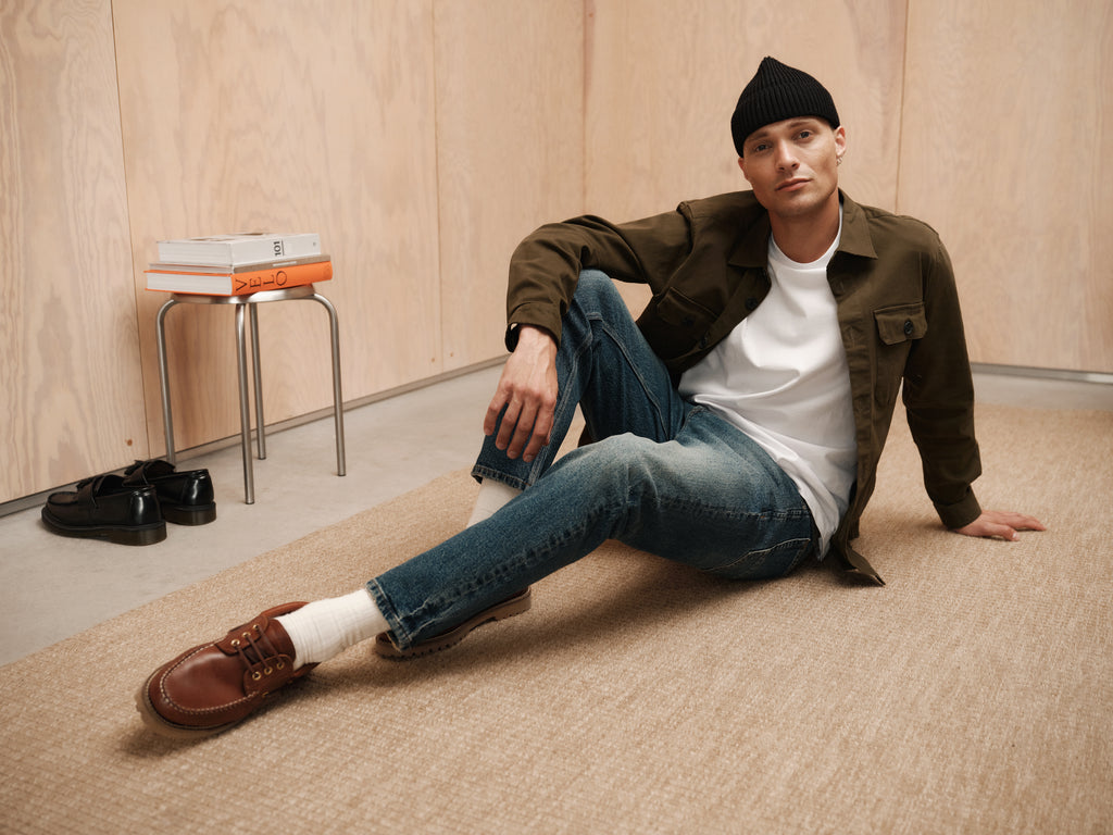 A man in a black beanie, olive jacket, white t-shirt, and brown shoes sits on a beige rug in a minimalist room, wearing Gabba’s Carl Oskin - Dark Blue Denim jeans for an effortlessly casual look.