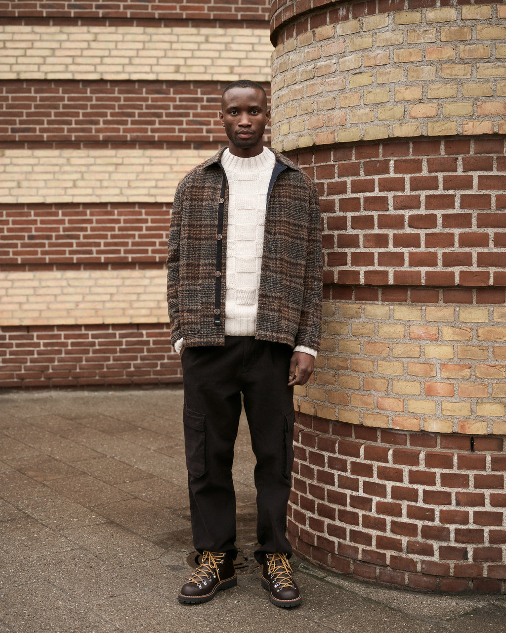 A man stands against a brick wall, wearing the Gabba Oscar J Check – Multi Check wool blend jacket over a white cable-knit sweater, with black pants and brown lace-up boots. He faces the camera with a neutral expression.
