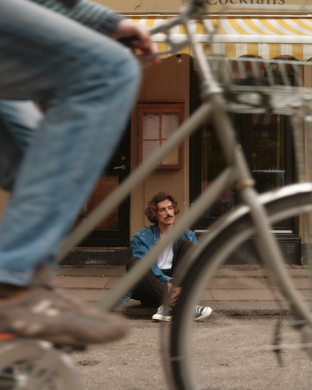 A person with curly hair sits on the sidewalk before a building with a striped awning, wearing Gabbas Topgun Jash - Black pants made from ripstop fabric. They gaze into the distance as a bicycle, partially obscuring them, passes by in the foreground.