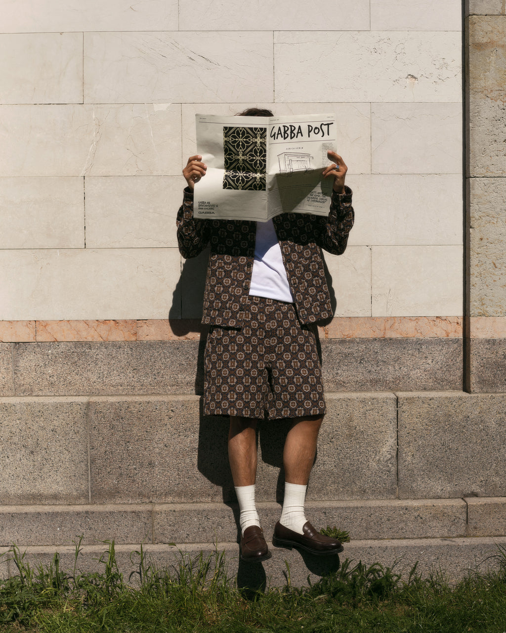 A person in a statement outfit, featuring Gabbas Fede Box Star set with boxy shorts and a matching jacket, reads the newspaper against a stone wall. The Gabba Post obscures their face as they pair white socks with brown loafers, grounding the loose look on grass.