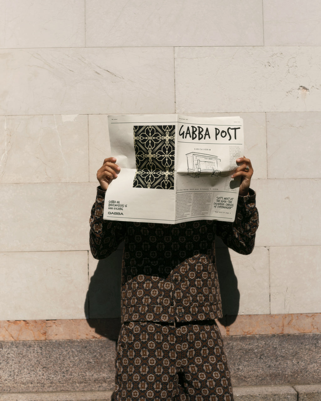A person wearing the Fede Box Star - Multi Pattern outfit by Gabba stands against a light stone wall, holding the Gabba Post newspaper to conceal their face. Their distinct style pops with the boxy shorts against the subtly textured backdrop.