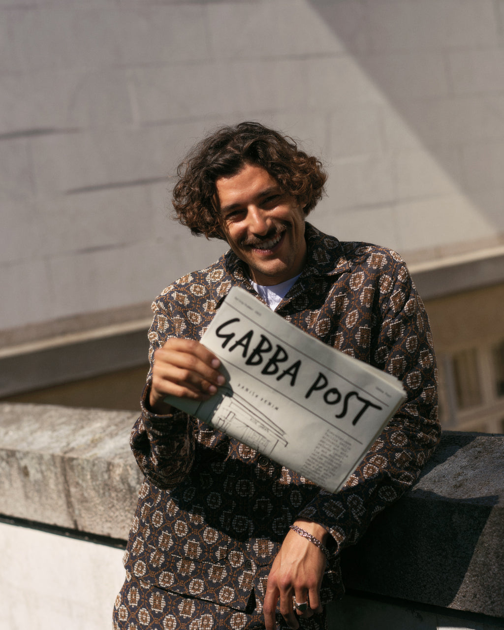 A smiling person with curly hair, dressed in a statement Fede Box Star - Multi Pattern outfit from Gabba, leans against a stone ledge. Theyre holding up a newspaper titled Gabba Post, with a blurred stone wall in the background.