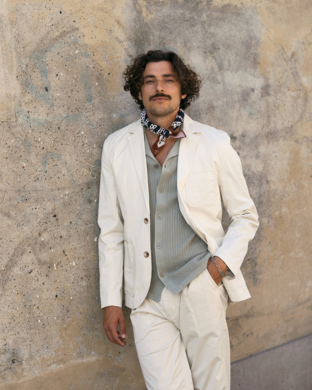 A person with curly hair and a mustache leans against a textured concrete wall, sporting the Josh Unite - Beige blazer by Gabba in a regular fit, paired with a buttoned-up cotton stretch shirt and an elegantly draped patterned scarf.