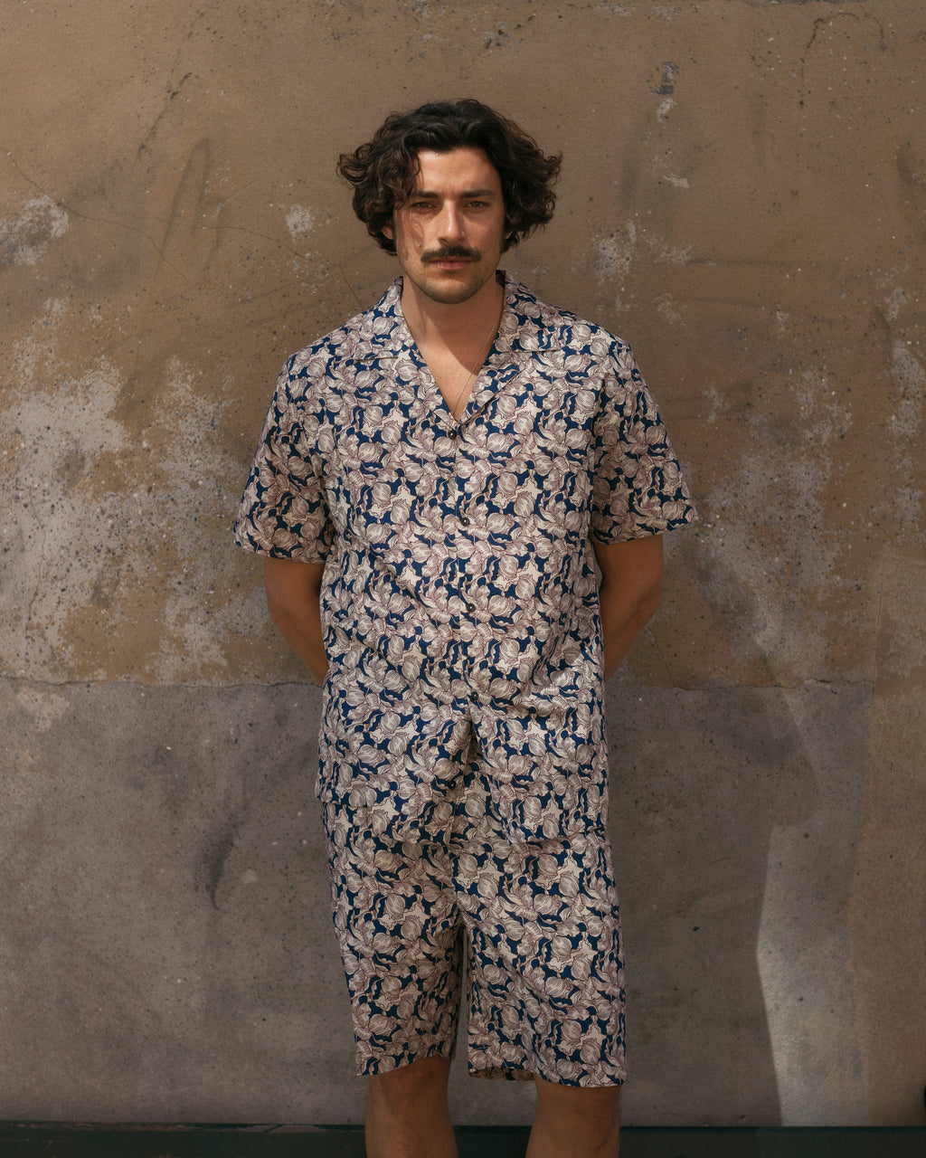A person with curly hair and a mustache stands against a textured brown and grey wall, wearing the Gabba Porter Epo - Blue Pattern matching short-sleeve shirt and premium shorts set, hands in pockets.