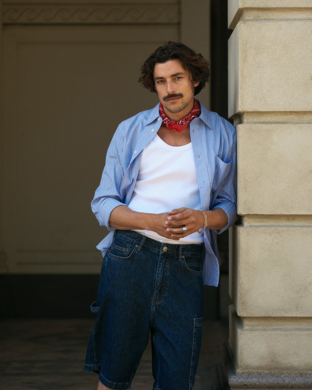 A curly-haired man with a mustache leans against a pillar, dressed in Gabbas Ugo Cargo Fone - Mid Blue Denim shorts. He wears a light blue shirt over a white tank top with a red bandana around his neck, exuding a relaxed and casual vibe that matches the loose fit of his outfit perfectly.