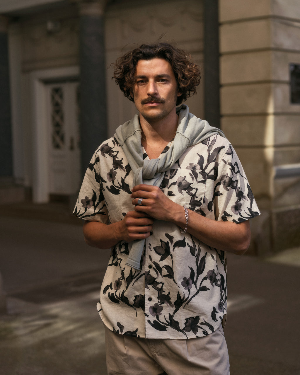 A person with curly hair and a mustache stands in a street wearing the Sven Reg Resort Bloom SS - Meteorite by Gabba with a light sweater over their shoulders, against a backdrop featuring columns and a partially visible door.