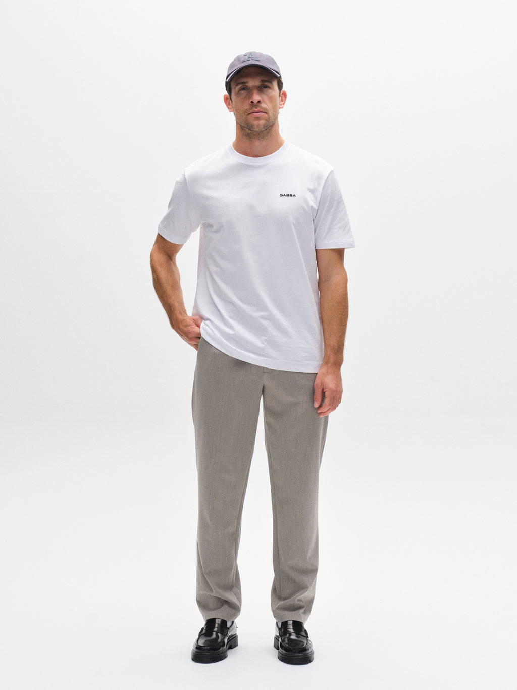 A man stands against a white background, wearing the Gabba GABdune Logo SS GOTS t-shirt in white, a light gray baseball cap, beige pants, and black shoes. One arm is bent with his hand on his hip.