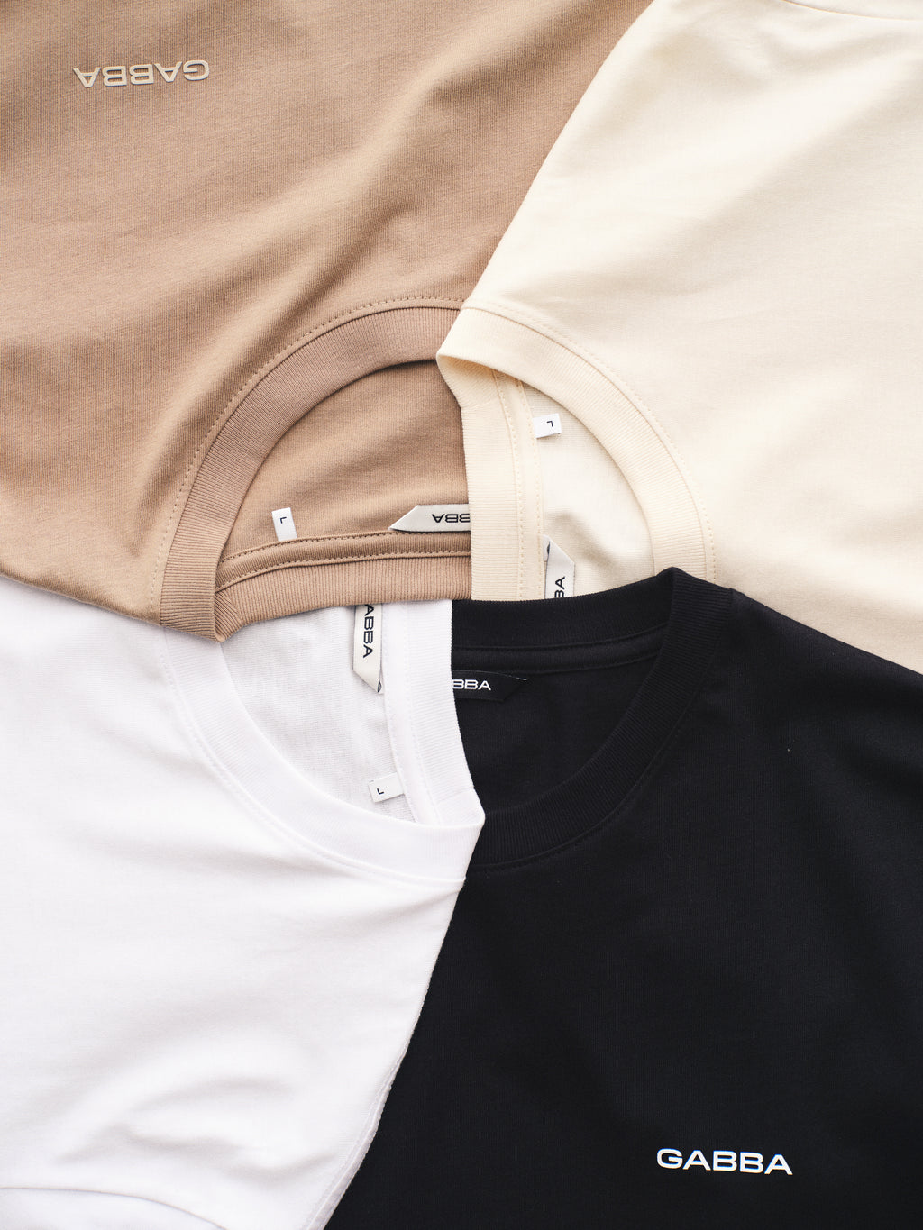 Four classic fit GABdune Logo SS GOTS t-shirts by Gabba in white, beige, cream, and black lay in a circle. Made from organic cotton, each features a visible neck label and the Gabba logo printed on the front.