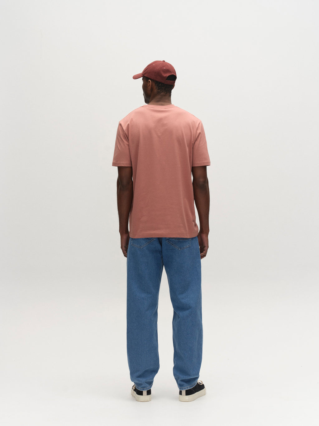 A person faces away from the camera, wearing a maroon cap, loose blue jeans, black-and-white sneakers, and the Gabba GABdune Logo SS GOTS t-shirt in Wood Pink, set against a plain light background.