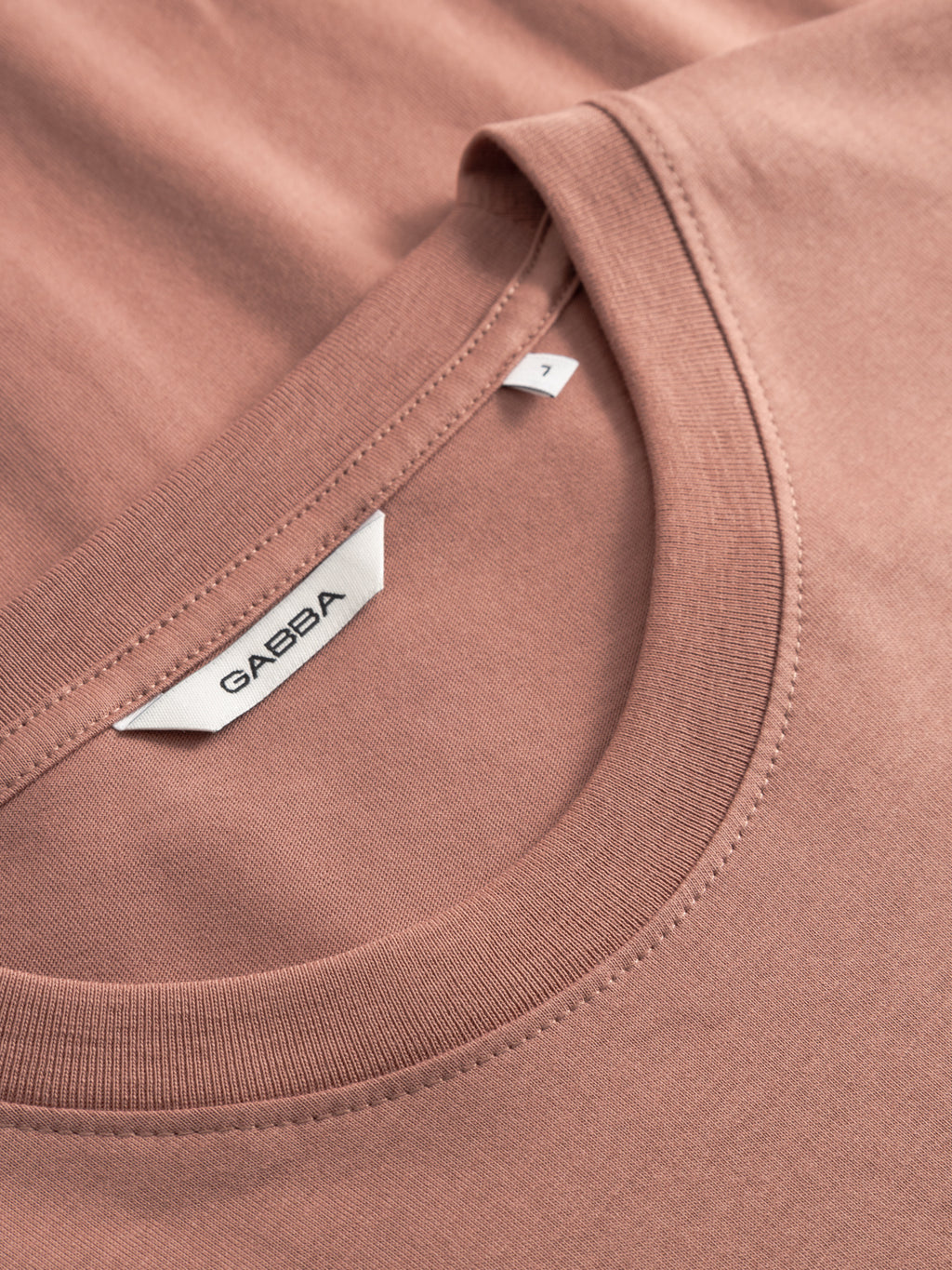 Close-up of a Gabba GABdune Logo SS GOTS T-shirt in Wood Pink, featuring a neatly stitched neckline and white label with Gabba branding and size M. The organic cotton fabric appears soft and smooth.