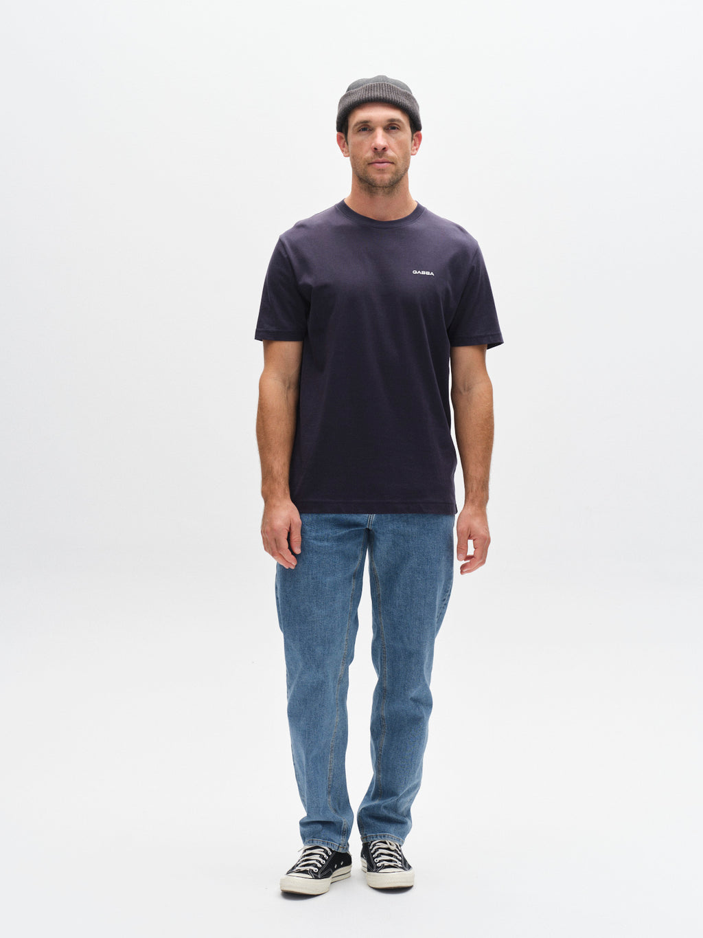 A man stands facing forward against a white background, wearing a gray beanie, blue jeans, black-and-white sneakers, and the Gabba GABdune Logo SS GOTS t-shirt in Total Eclipse. His arms rest at his sides.