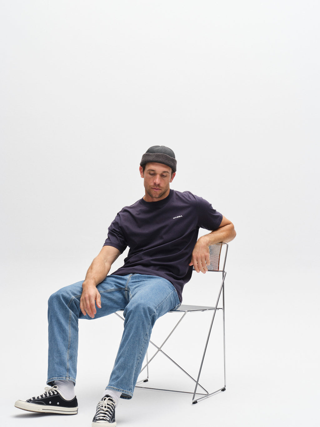 A man in a black beanie, Gabba GABdune Logo SS GOTS t-shirt in Total Eclipse, blue jeans, and black Converse sneakers sits casually on a metal chair against a plain white background.