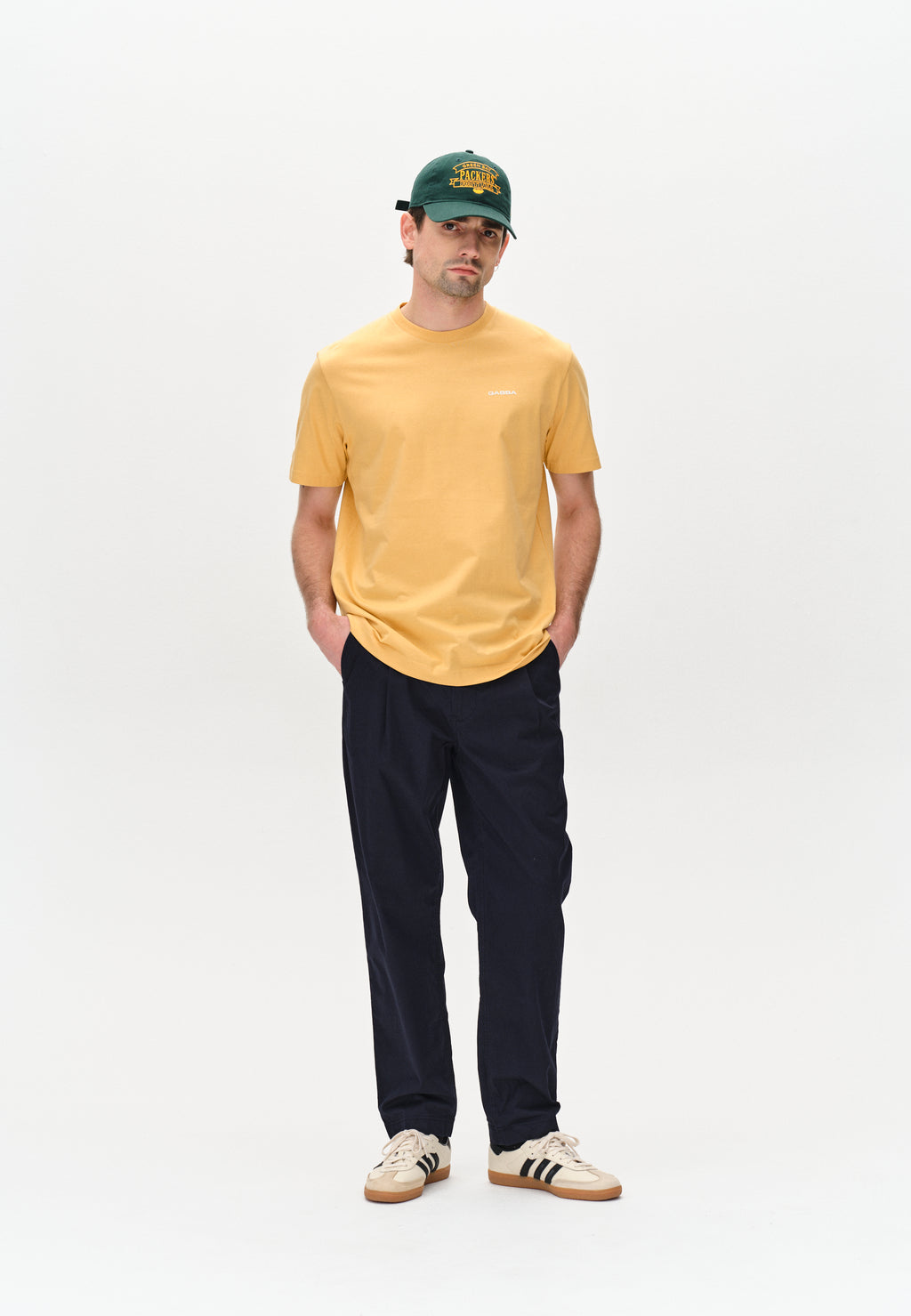 A person wears a Gabba Dune Logo SS GOTS in Yellow Jojoba, black pants, and white sneakers with black stripes. Hands are in their pockets as they sport a dark green baseball cap against a plain background.