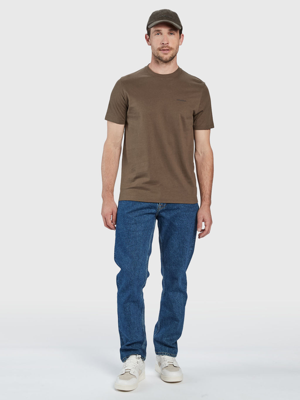 A man stands facing forward, wearing the Gabba GABdune Logo SS GOTS t-shirt in Crocodile, blue jeans, white sneakers, and a brown cap against a plain white background.