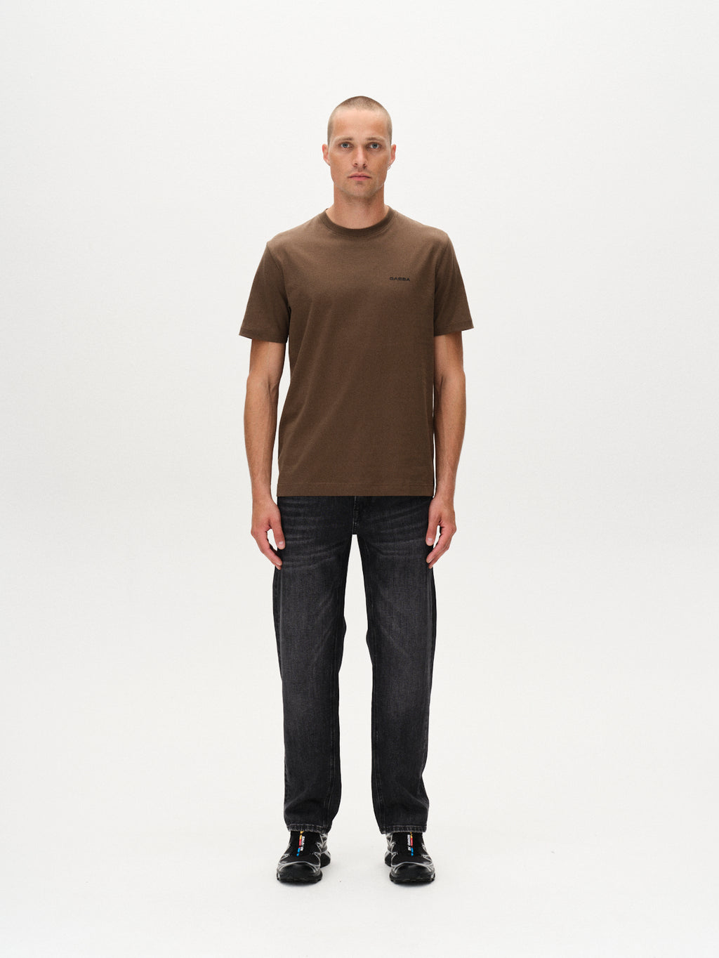 A man with a buzz cut faces forward against a plain white background, wearing the Gabba Dune Logo SS GOTS - Crocodile t-shirt in brown, dark jeans, and black sneakers.