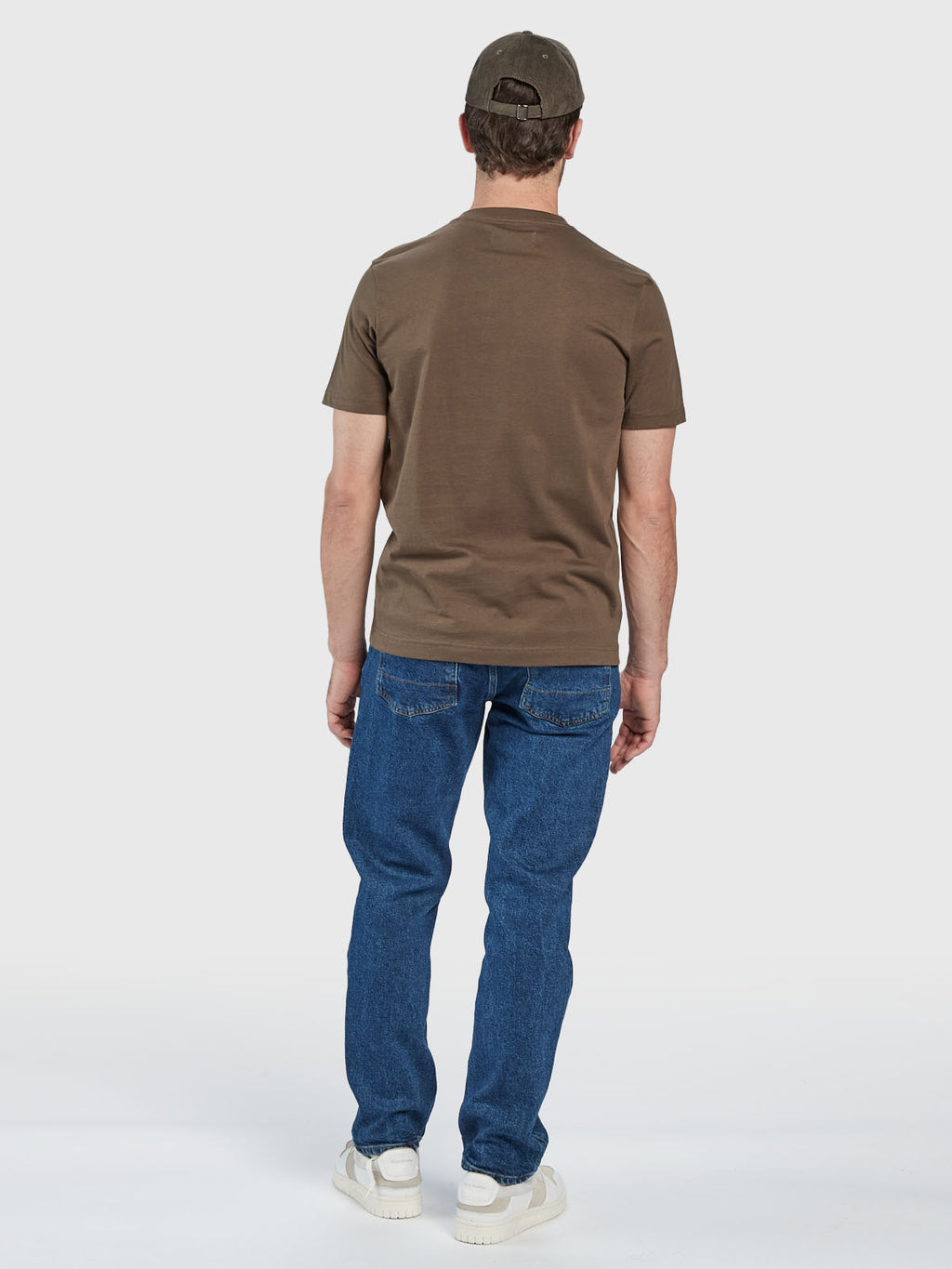 A man faces away, wearing the Gabba GABdune Logo SS GOTS - Crocodile t-shirt in brown, paired with blue jeans, white sneakers, and a matching brown cap against a plain light background.