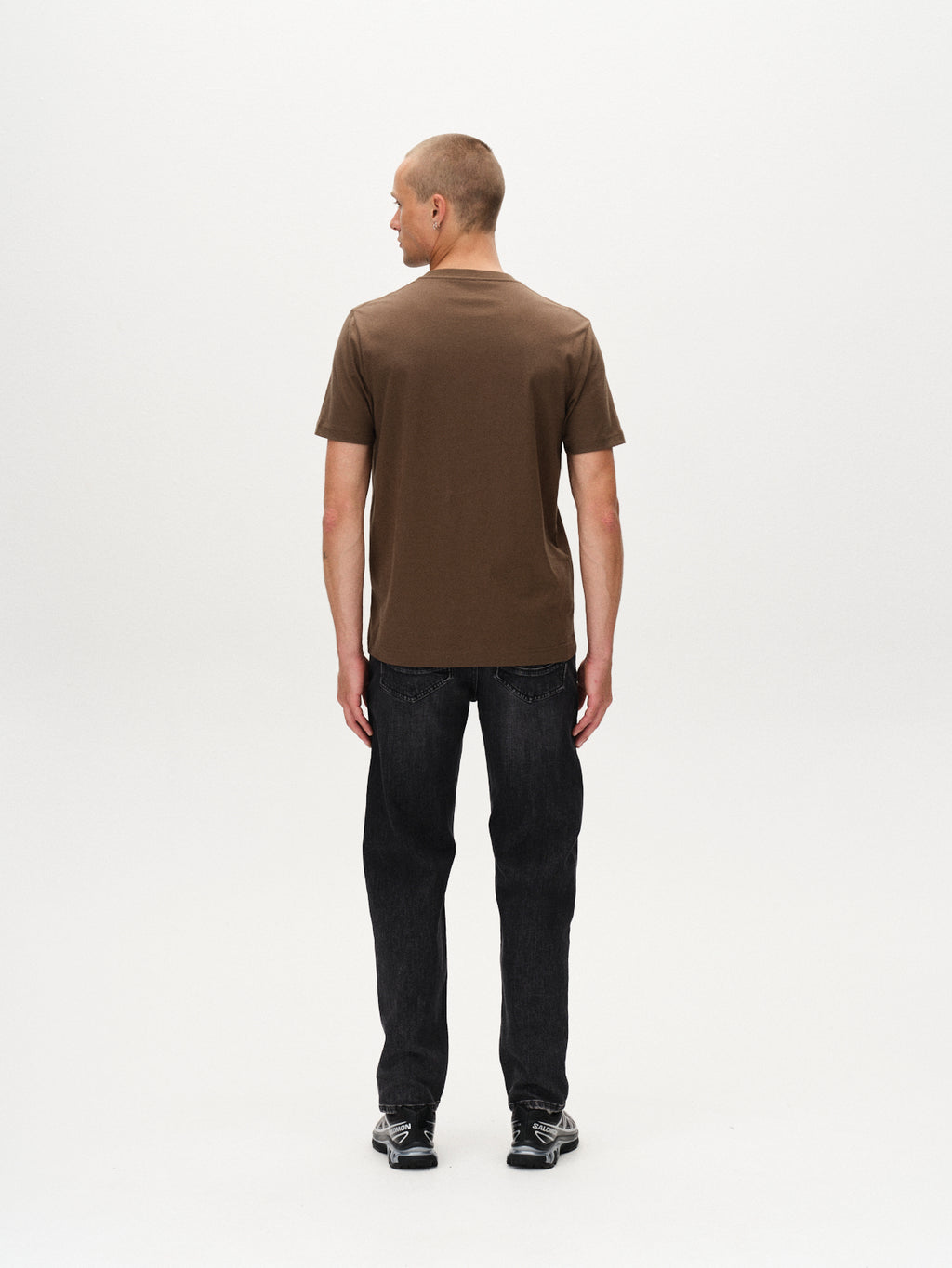 A man with a buzz cut, seen from behind, wears the Gabba Dune Logo SS GOTS - Crocodile brown organic cotton t-shirt, paired with black jeans and sneakers, standing against a plain white background.