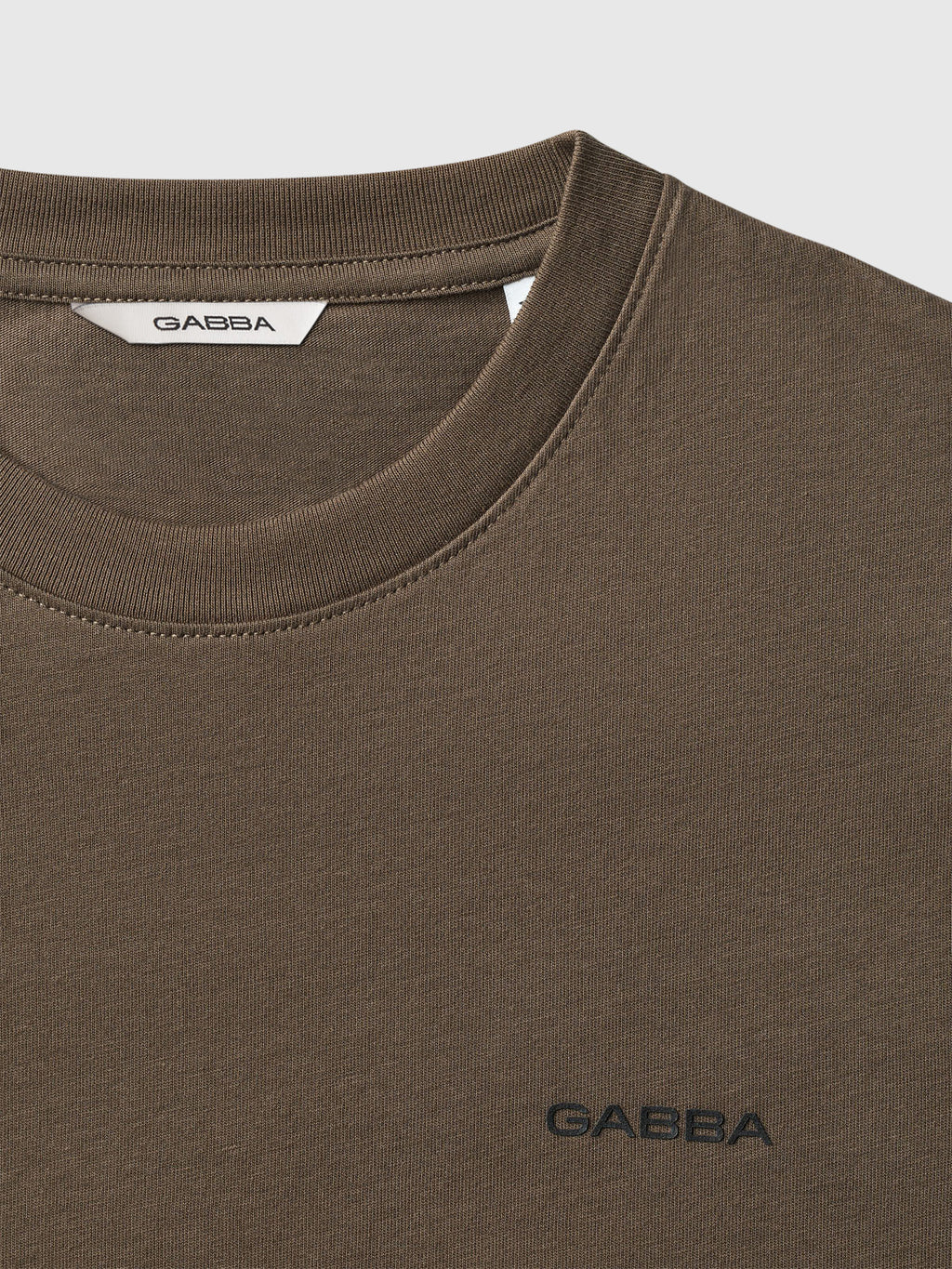 Close-up of a brown organic cotton t-shirt, the Gabba Dune Logo SS GOTS - Crocodile, featuring Gabba stitched in black on the chest and visible on the white neck tag.