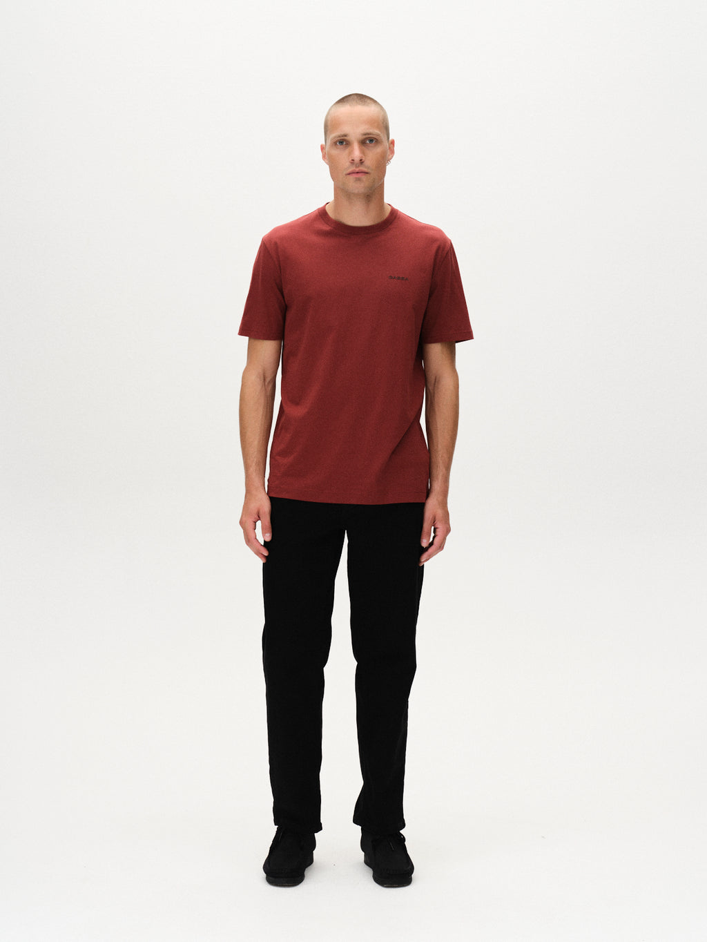 A person stands facing forward against a plain white background, wearing the Gabba Dune Logo SS GOTS t-shirt in Red Russet, paired with black pants and black shoes.