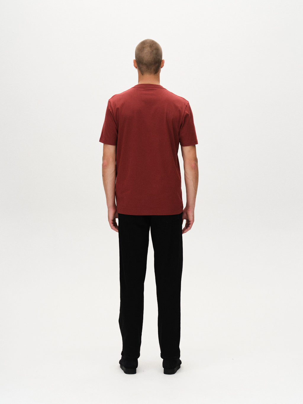 A person with short hair faces away from the camera, wearing Gabbas Dune Logo SS GOTS t-shirt in Red Russet, paired with black pants and shoes, against a plain white background.