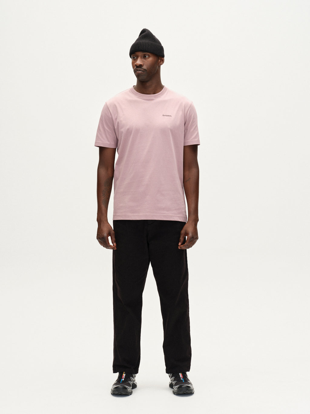 A man in the Gabba Dune Logo SS GOTS t-shirt in Dust Purple stands against a plain white background, wearing a black beanie, black pants, and black sneakers. He looks slightly left with a neutral expression.