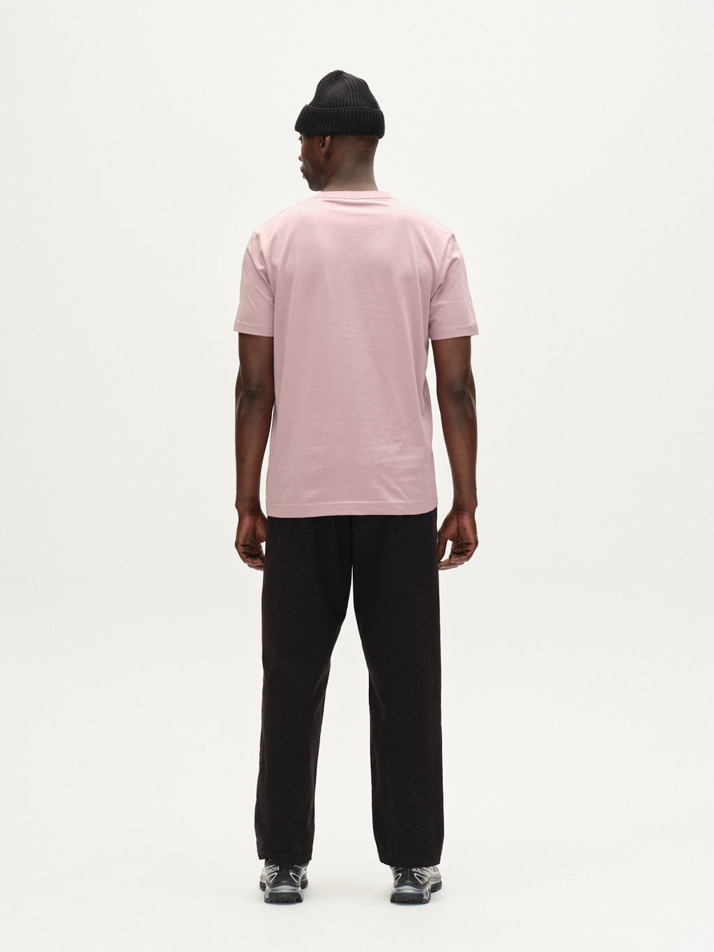 A man faces away from the camera, wearing the Gabba Dune Logo SS GOTS tee in Dust Purple, black pants, black sneakers, and a black knit beanie against a plain white background.