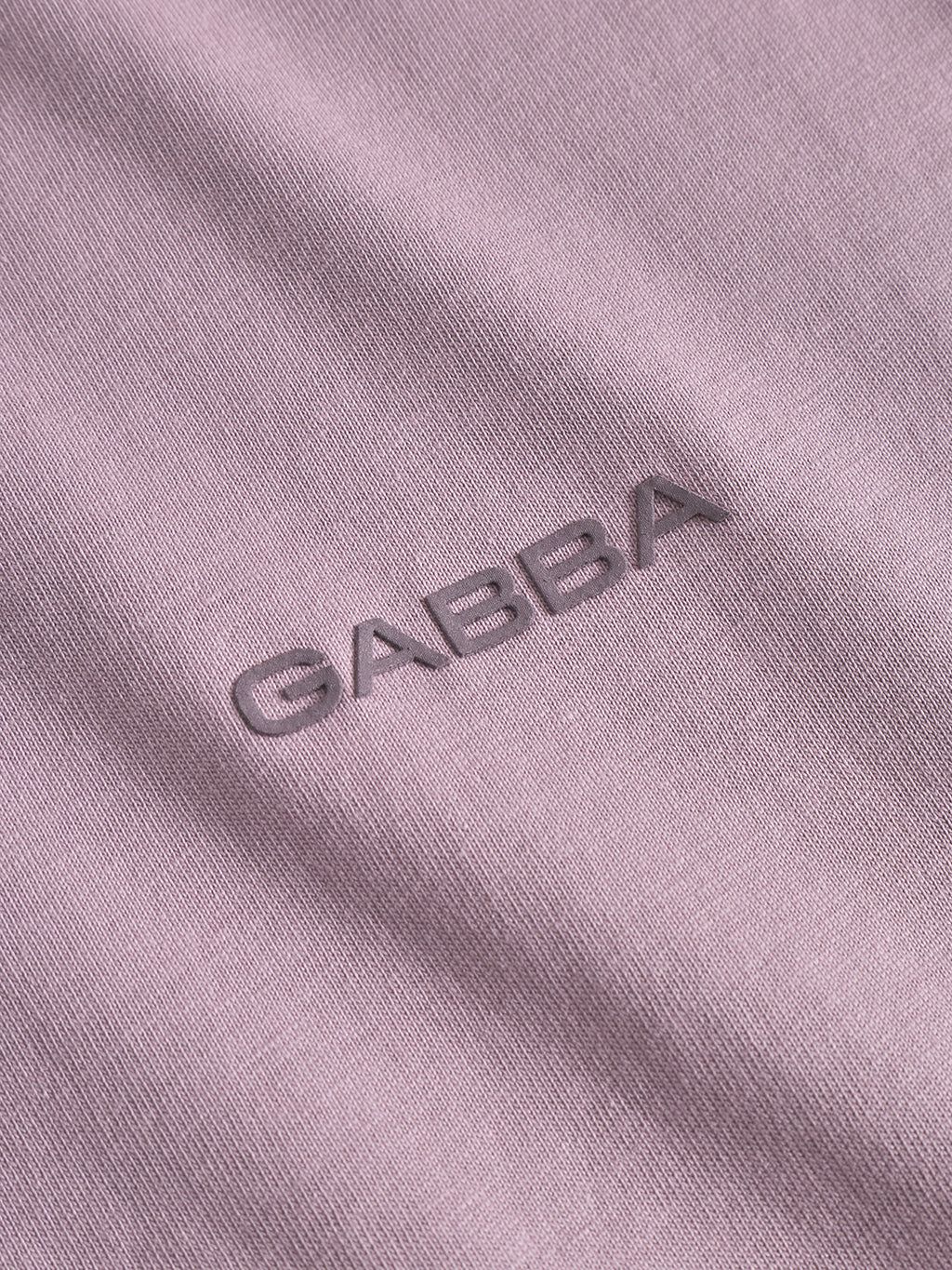 Close-up of Gabba’s Dune Logo SS GOTS t-shirt in Dust Purple, showing organic cotton fabric with “GABBA” embroidered in matching thread at the center. The texture and shadows highlight this premium basic.