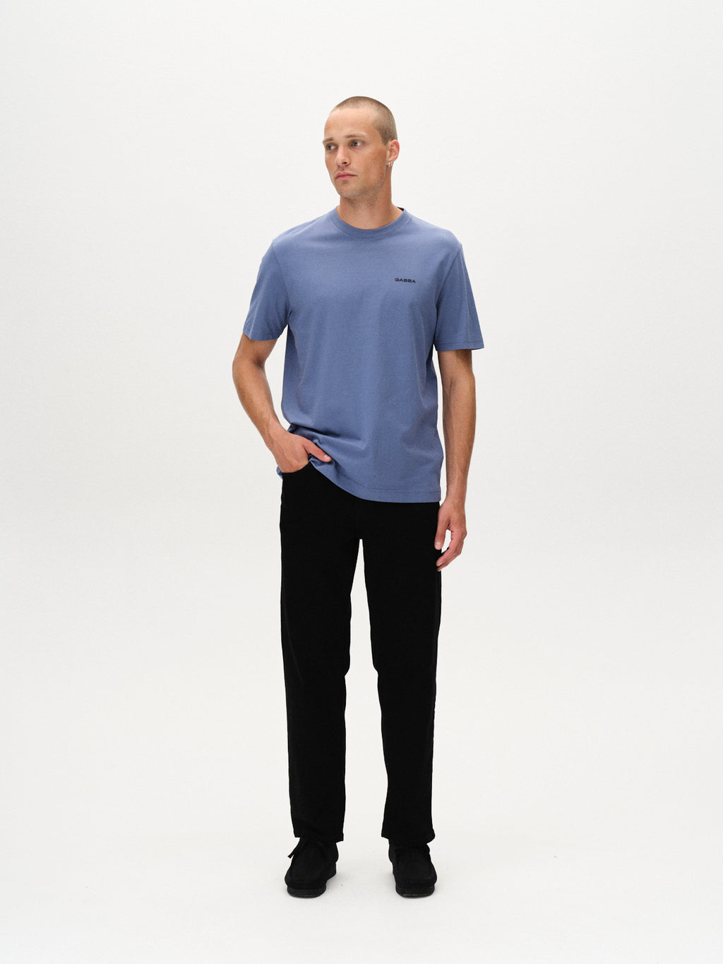 A man with short hair stands against a white background, wearing Gabbas Dune Logo SS GOTS - Blue Horizon t-shirt made from GOTS-certified organic cotton, paired with black pants and shoes. One hand is in his pocket as he looks to the side.