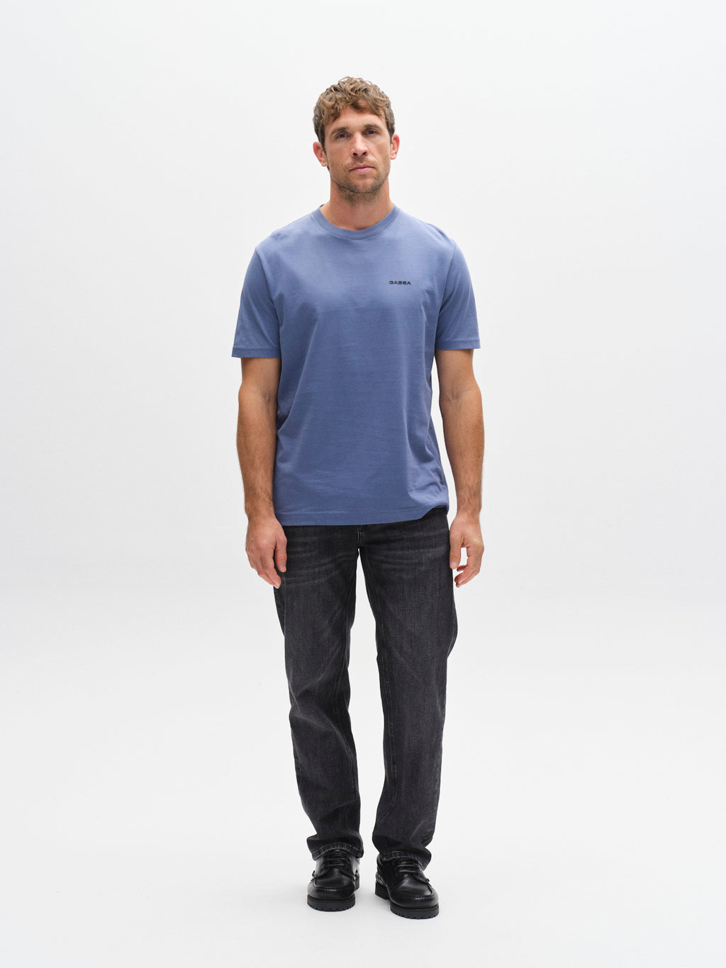 A man stands facing forward against a white background, wearing the Gabba GABdune Logo SS GOTS t-shirt in Blue Horizon, paired with black jeans and shoes. His expression is neutral and his arms are relaxed by his sides.