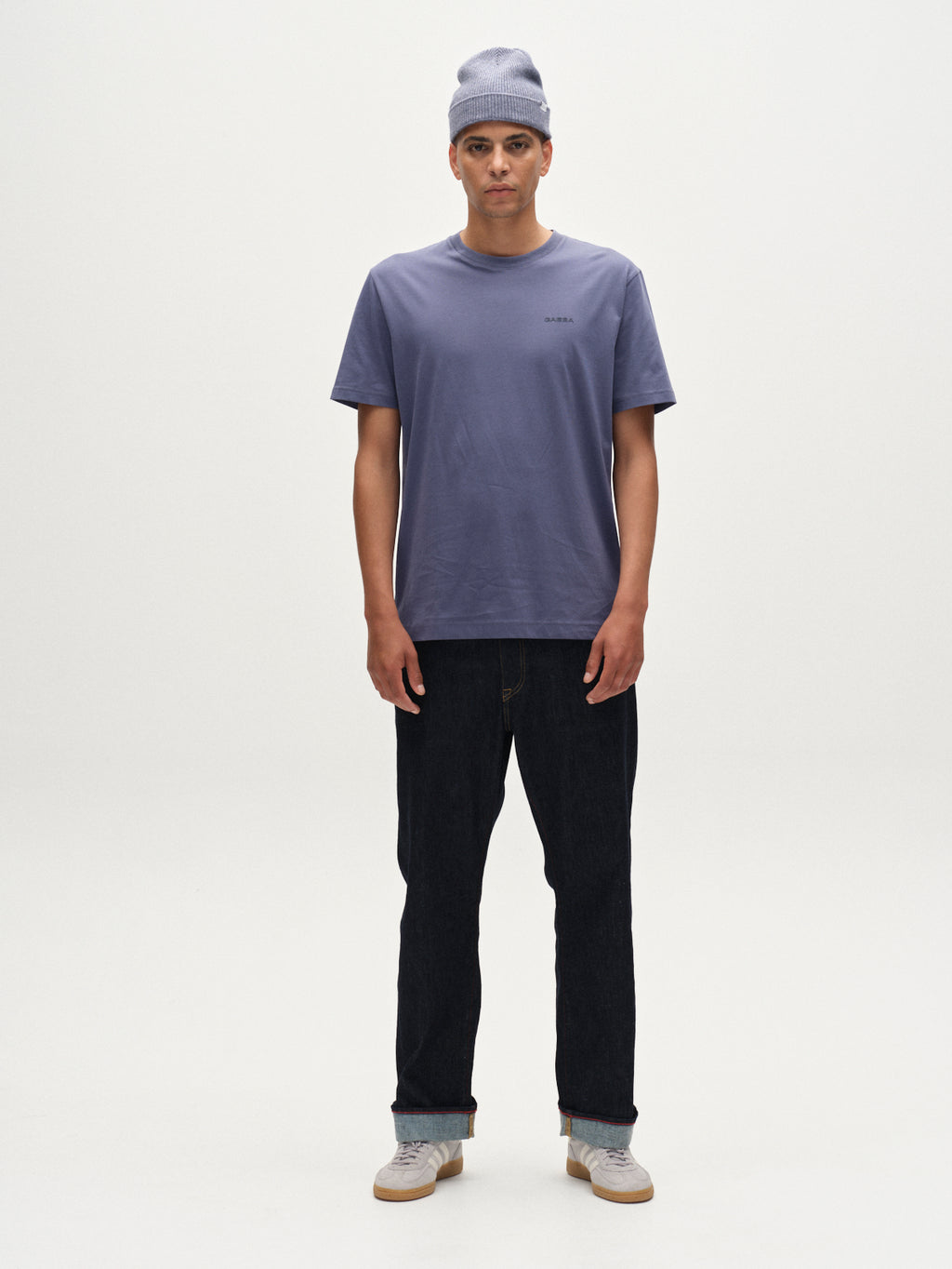 A person stands facing forward against a plain white background, wearing the Gabba Dune Logo SS GOTS t-shirt in Vintage Indigo, a matching beanie, dark jeans, and light-colored sneakers.