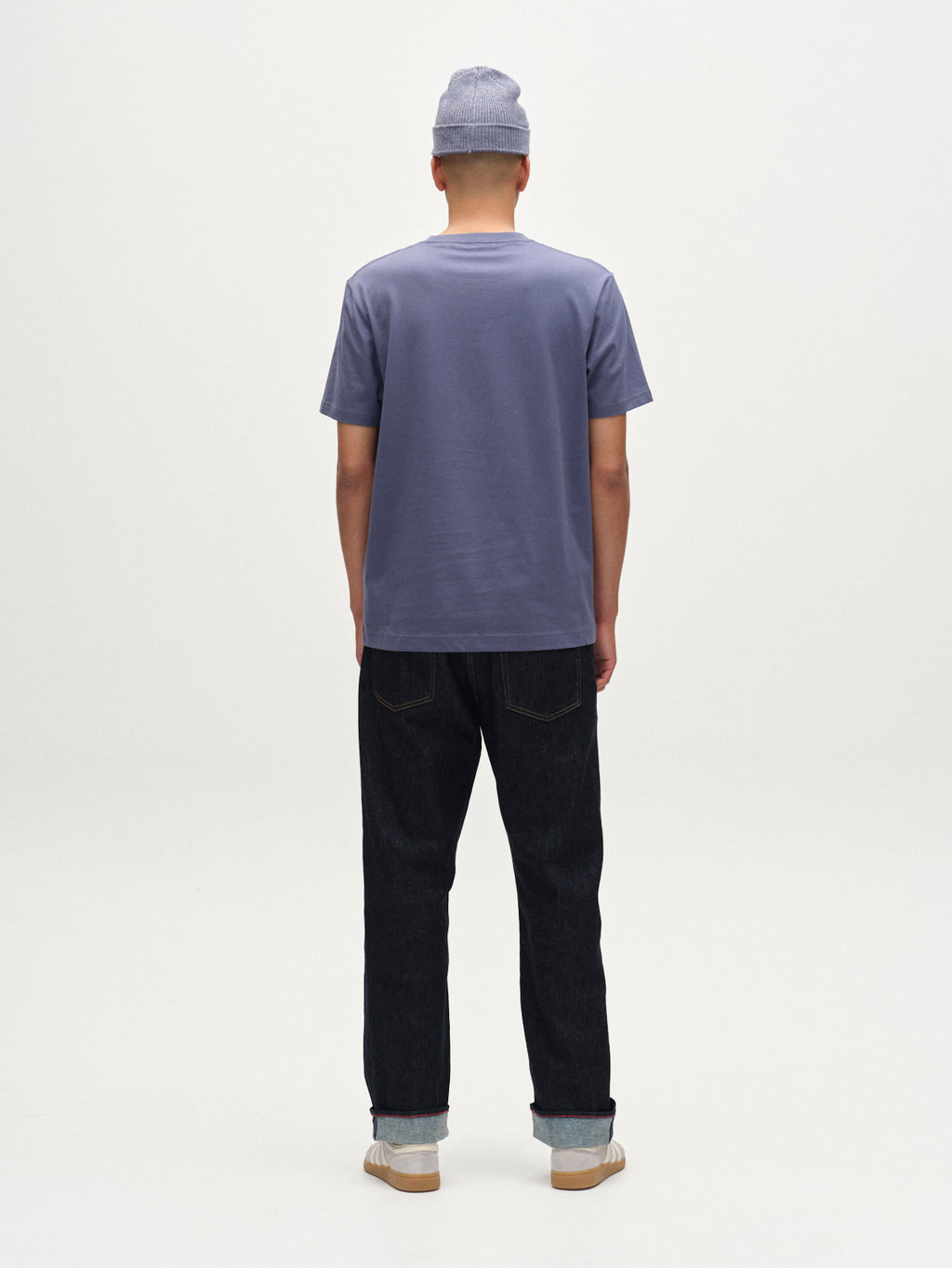 A person stands facing away from the camera against a plain white background, wearing Gabba’s Dune Logo SS GOTS t-shirt in Vintage Indigo with dark cuffed jeans, a light gray beanie, and white sneakers.