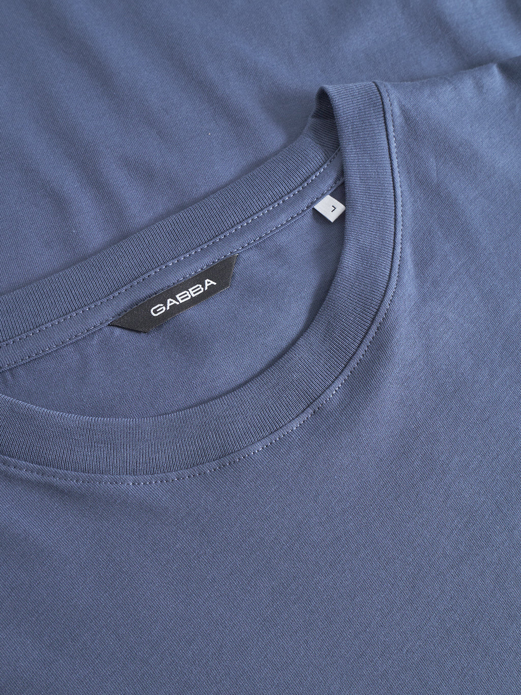 A close-up of two overlapping Dune Logo SS GOTS - Vintage Indigo t-shirts by Gabba, with the top shirt’s brand label and size tag visible at the collar and its soft, smooth fabric on display.