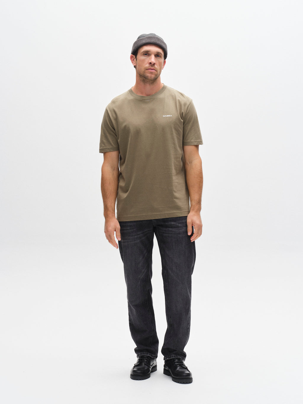 A man wears a Gabba GABdune Logo SS GOTS t-shirt in Deep Lichen Green with a gray beanie, dark jeans, and black shoes, standing against a plain white background and looking straight ahead with arms relaxed at his sides.