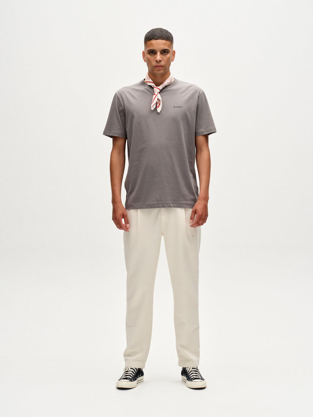 A man faces forward against a plain white background, wearing the Gabba Dune Logo SS GOTS t-shirt in dust sage green, cream pants, black-and-white sneakers, and a red-and-white scarf tied around his neck.