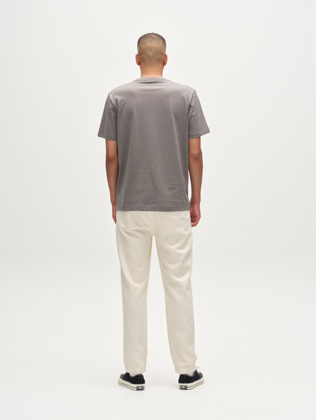 A person stands facing away from the camera against a plain white background, wearing the Gabba Dune Logo SS GOTS t-shirt in Dust Sage Green, off-white pants, and black shoes with white soles.