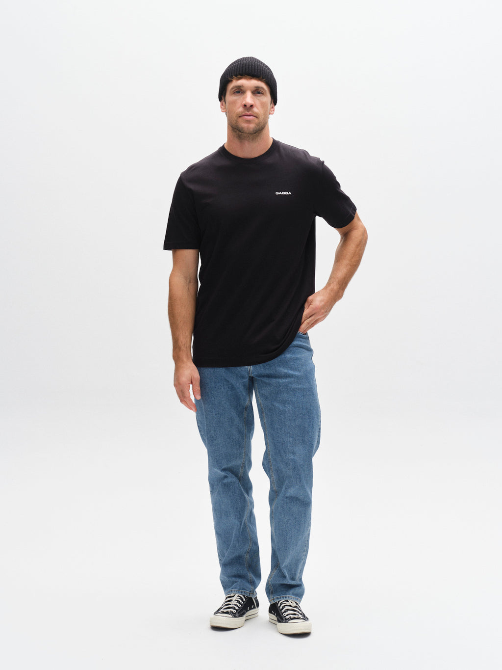A man wearing the Gabba GABdune Logo SS GOTS black organic cotton t-shirt stands against a plain white background, paired with a black beanie, blue jeans, and black sneakers, one hand resting on his hip.