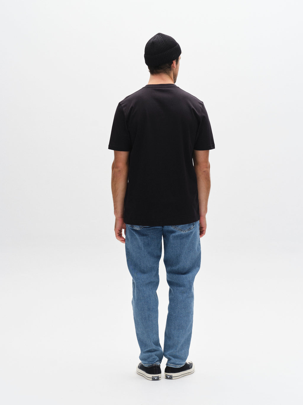 A man faces away from the camera, wearing a Gabba GABdune Logo SS GOTS black t-shirt, blue jeans, a black beanie, and black and white sneakers against a plain white background.