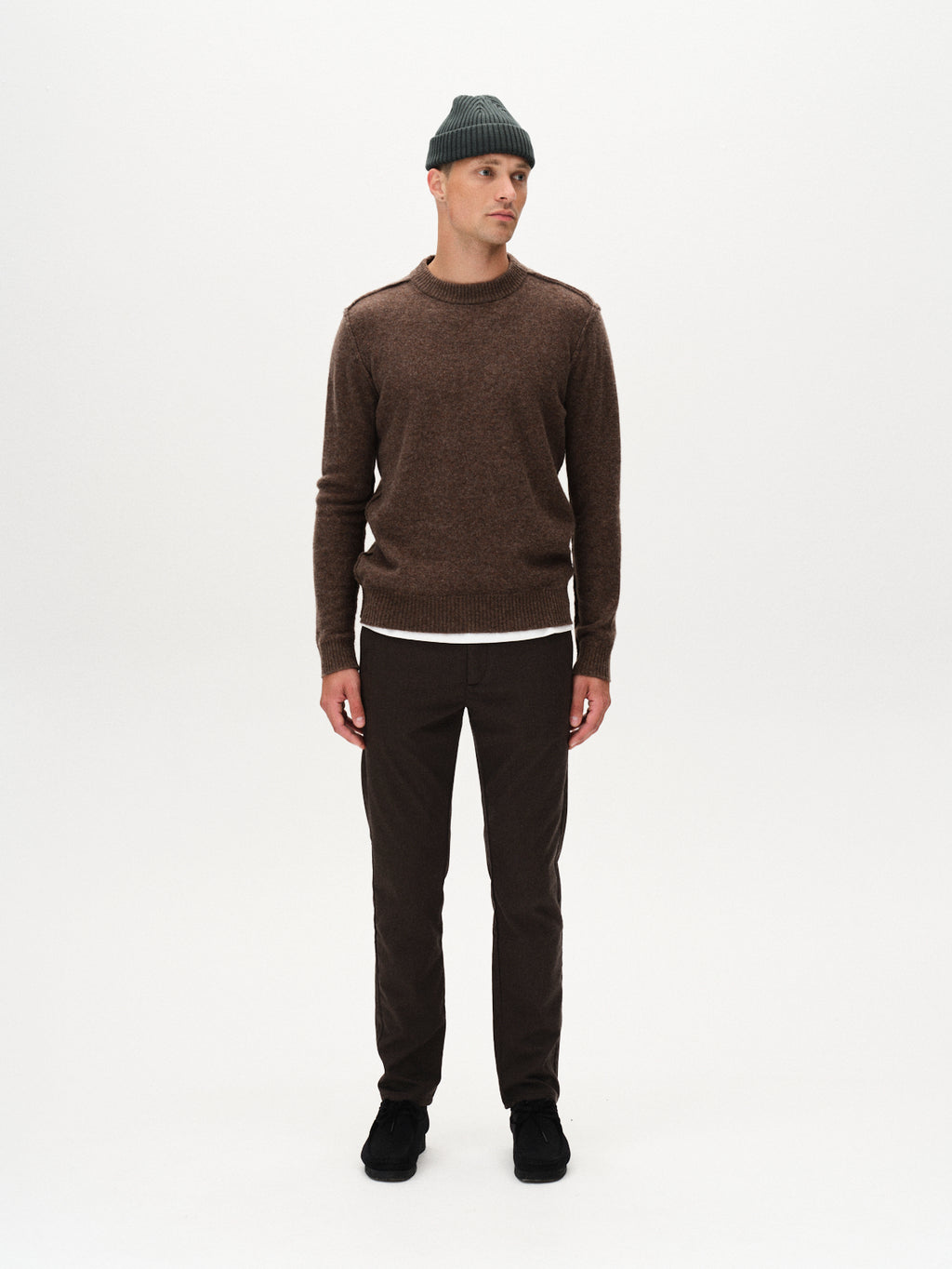 A man models the Gabba Paul Domo - Dk. Brown sweater, styled with a gray beanie, white undershirt, slim-fit chinos with French pockets, and black shoes. He stands against a white background, glancing to the side with relaxed hands.